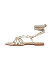 Sandalias planas metálicas doradas con múltiples tiras, detalle de envoltura en el tobillo y una suela beige. Diseño de punta abierta con una estructura minimalista.