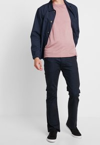 Chaqueta ligera de marina con botones dorados sobre una camisa rosa claro, combinada con jeans oscuros y zapatillas negras con suelas blancas.