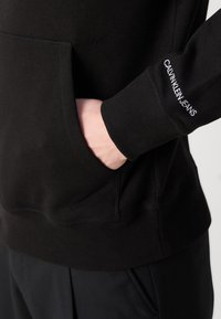 Käsi musta dressipluusi taskus, kus randmekoorel on tikitud Calvin Klein Jeansi logo.