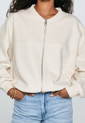 Nainen pukeutuneena kermaan vivahtavaan vetoketjulliseen collegepaitaan ja vaaleansinisiin farkkuihin, kädet farkkujen taskuissa ja vasemmassa ranteessa kultaiset rannekorut.