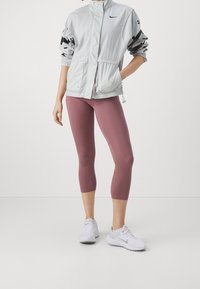 Persoon in een lichtgrijze Nike-jas, mauve cropped leggings en witte Nike hardloopschoenen, staand met één hand in de zak.