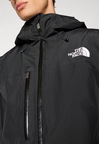 Giacca nera impermeabile con colletto alto, zip frontale e logo sul petto. Presenta un cappuccio regolabile e una trama di tessuto liscia e elegante.
