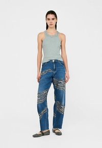 Canotta grigio chiaro con scollatura decorativa, abbinata a jeans blu con motivi testurizzati beige e bianchi. Scarpe piatte nere completano il look.