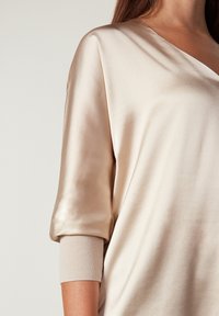 Satin beige topp med V-ringning, lös passform och uppvikta ärmar som har en mjuk stickad manschett. Slät textur med subtil glans.