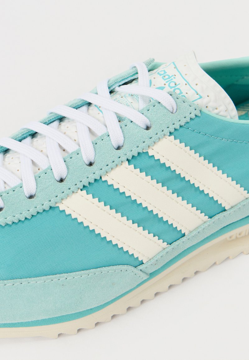 Zapatilla deportiva turquesa con acentos blancos, con tres franjas blancas, malla texturizada y un diseño de suela estriada. Cuello ligeramente acolchado.