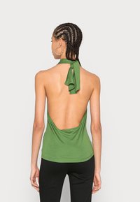 Top halter verde com design de costas abertas, tecido suave e um laço amarrado ao pescoço. Usado com calças pretas.