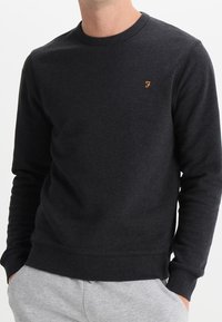 Sudadera gris oscuro de una tela suave y texturizada, con cuello redondo y puños acanalados. Presenta un pequeño logo naranja en el pecho.