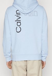 Felpa blu chiaro realizzata in tessuto morbido con un grande logo nero "Calvin Klein" stampato verticalmente sulla schiena, dotata di cappuccio con cordoncino.