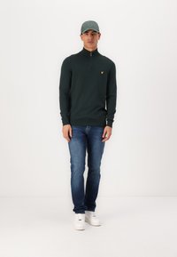 Pulover închidere cu fermoar de culoare verde închis, cu mâneci lungi, purtat cu blugi blue și teniși albi. Prezentă un logo mic galben pe piept.