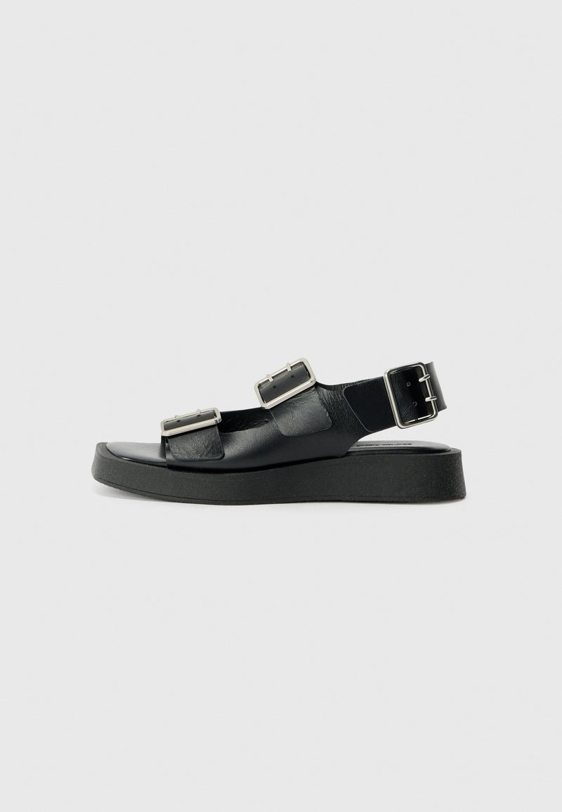 Μαύρες δερμάτινες σαγιονάρες με δύο λουριά με αγκράφες, επίπεδη σόλα και υφή. Ο σχεδιασμός είναι μινιμαλιστικός με φαρδύ τακούνι.