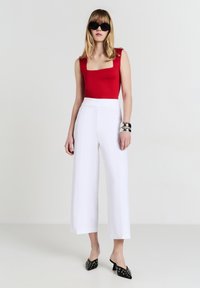 Body rosso con scollo a quadri, pantaloni bianchi a gamba larga, mule neri con borchie, bracciali d'argento, modella in posa su uno sfondo neutro.