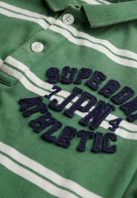 Zielona koszulka polo w białe paski, z haftowanym granatowym napisem "SUPER" i "ATHLETIC," z dwoma białymi zapięciami na guziki.