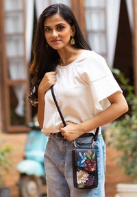 Schwarze Umhängetasche mit floralem Stickerei in Pink, Blau und Weiß; kombiniert mit einer weißen Bluse und hochgeschnittenen blauen Jeans.