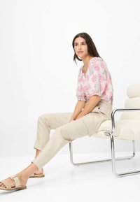 Bluse mit floralem Muster in Pink- und Blautönen, kurze Puffärmel; kombiniert mit beigen, taillierten Hosen und neutralen Slipper-Sandalen.