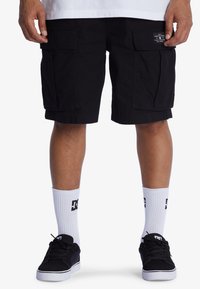 Schwarze Cargo-Shorts aus einem strapazierfähigen Stoff, mit Seitentaschen und geradem Schnitt. Kombiniert mit schwarzen Sneakers und weißen Knöchelsocken.