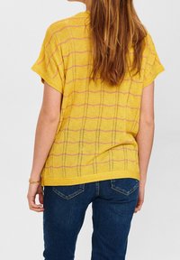 Gul stickad topp med korta ärmar, med ett texturerat vågmönster och rosa ränder. Matchad med mörkblå jeans, sedd bakifrån.