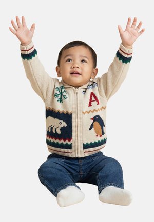 Tout-petit assis les bras levés, portant un pull à fermeture éclair beige avec des motifs d'animaux et de flocons de neige, un jean bleu et des chaussettes blanches.