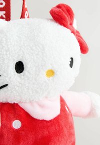 Mjuk Hello Kitty-leksak i vitt och rött. Mjukt, lurvigt material med ett runt huvud, röd klänning och en rosett. Gul näsa och svarta ögon.