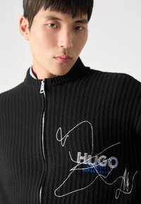 Svart ribbet glidelåsjakke med brodert "HUGO"-logo i blått og hvitt. Høy krage og glatt metallglidelåsdetalj.