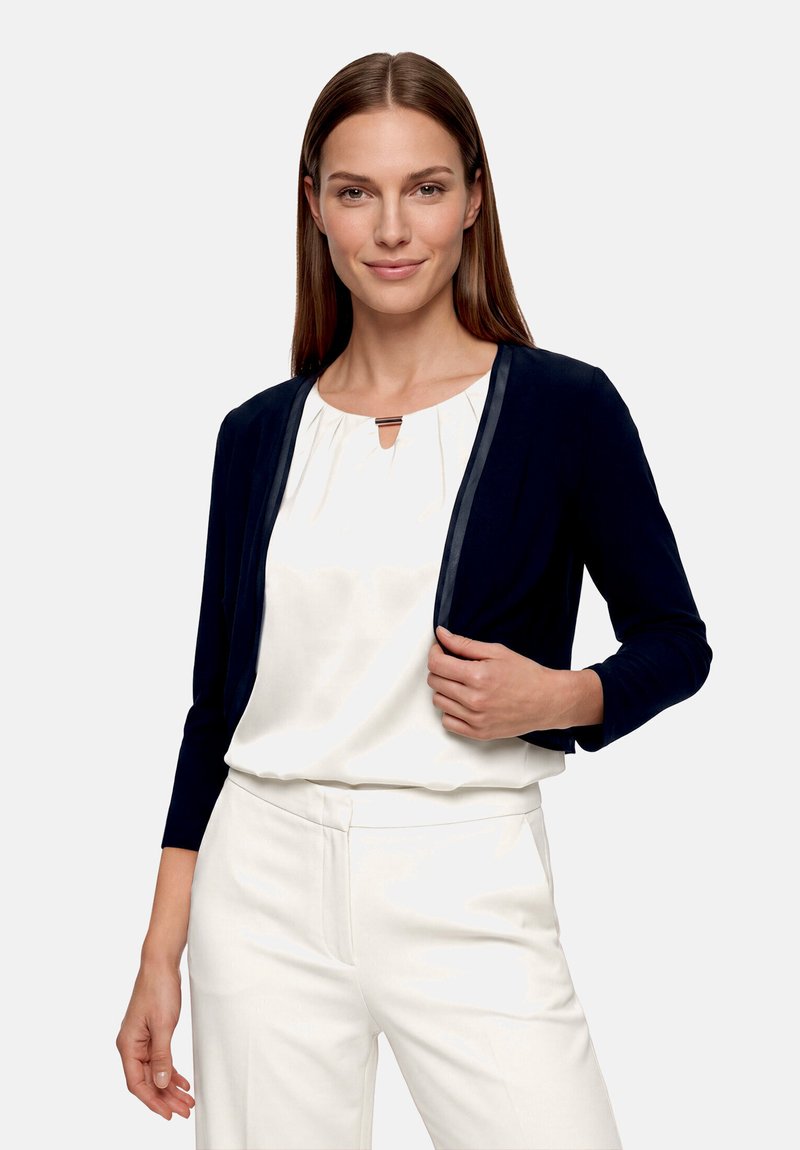 Vrouw draagt een witte blouse, witte broek en een donkerblauwe cropped jas, staand tegen een eenvoudige lichte achtergrond.