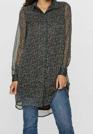 Femme portant une tunique florale transparente à manches longues et boutonnée sur un jean bleu, tenant l'ourlet de la tunique d'une main.