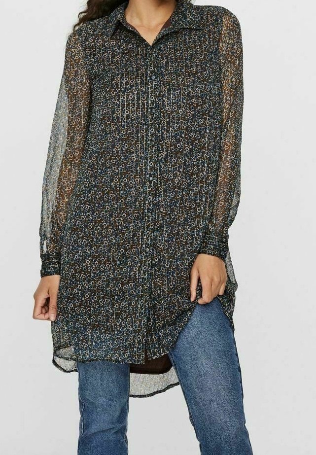 Femme portant une tunique florale transparente à manches longues et boutonnée sur un jean bleu, tenant l'ourlet de la tunique d'une main.