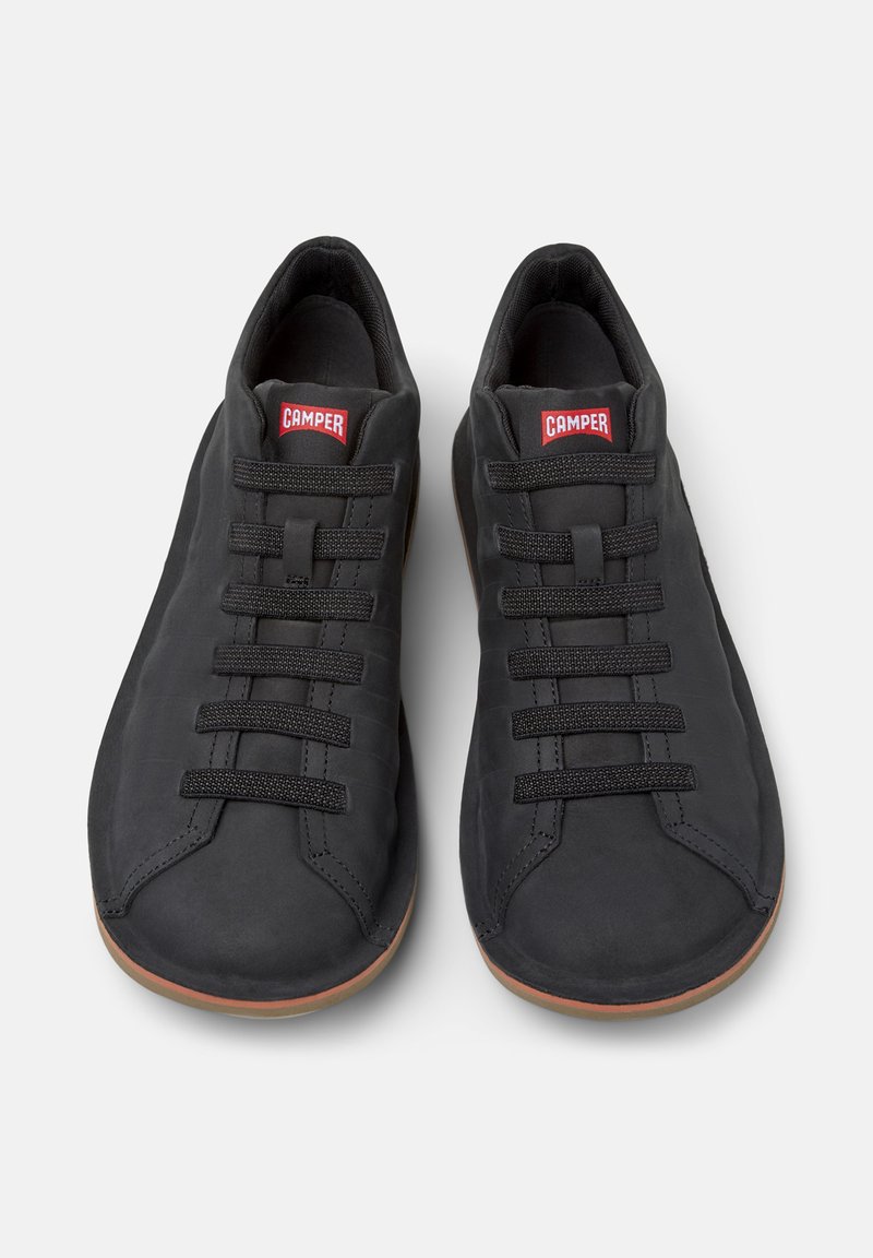Chaussures noires décontractées avec des lacets élastiques et une semelle en caoutchouc, arborant un logo rouge Camper sur la languette, sur un fond neutre.