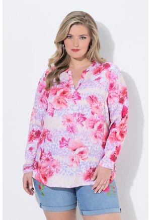 Frau mit blondem Haar, die eine rosa geblümte Langarmbluse und blaue Jeansshorts mit bestickten Blumen trägt, steht vor einem weißen Hintergrund.