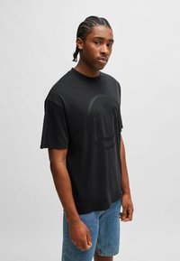 Sort bomulds t-shirt med et stort sort smiley ansigt grafik på forsiden. Korte ærmer, afslappet pasform. Bæret med blå denim shorts.