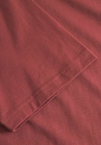 Vinrød bomulds-T-shirt med en glat tekstur, der har en detaljeret syet kant. Stoffet ser let krøllet ud, hvilket indikerer blødhed.