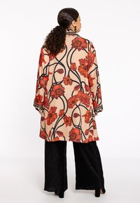 Beige farbener, floraler langer Mantel mit roten Blumen und schwarzen spiralförmigen Linien. Er hat lange Ärmel und dezente Bündchen und wird mit schwarzen Hosen kombiniert.