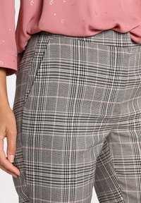 Un pantalon à carreaux gris présente un mélange de lignes noires et roses, conçu avec deux poches latérales et une coupe ajustée. Texture douce visible.