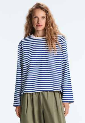 Mladá žena s dlhými vlnitými vlasmi, oblečená v modro-bielej pruhovanej dlhorukávovej košeli a voľných olivovo zelených nohaviciach, stojaca pred jednofarebným pozadím.