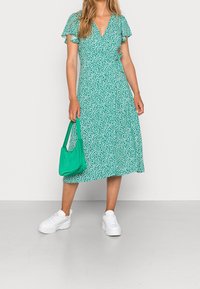 Grünes gemustertes Wickelkleid mit kurzen Rüschärmeln, kombiniert mit einer grünen Handtasche und weißen klobigen Sneakers. Mittellange Silhouette.