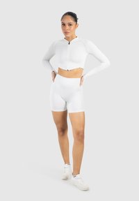 Haut de sport à manches longues blanches, coupé, avec une fermeture éclair sur le devant, associé à un short blanc taille haute. Tissu lisse, design ajusté.