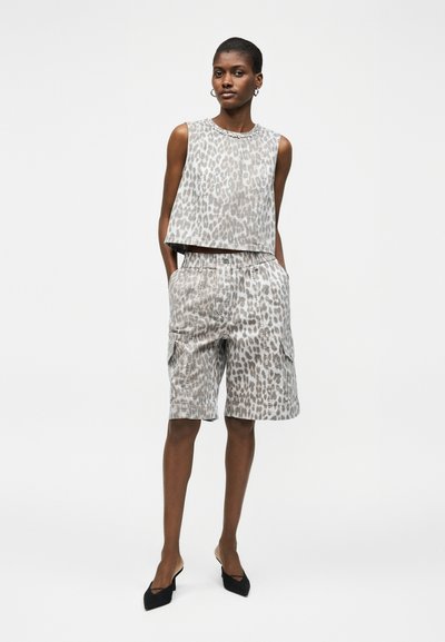 Γυναίκα στέκεται με τα χέρια στις τσέπες, φοράει αμάνικη μπλούζα με λεοπάρ σχέδιο και ταιριαστό μακρύ σορτς, σε συνδυασμό με μαύρα μυτερά τακούνια.