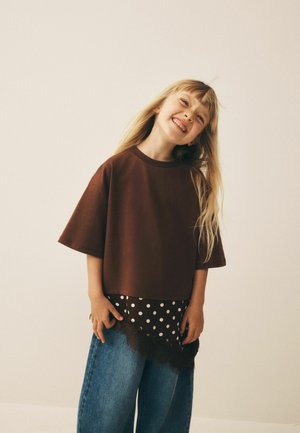 Giovane ragazza sorridente inclina la testa indossando una maglietta marrone oversize, gonna a strati con pois e jeans larghi blu su sfondo beige.