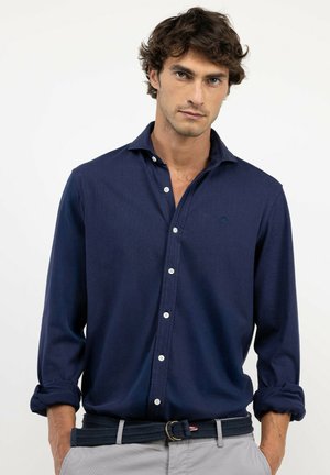 Hombre con una camisa azul oscuro abotonada con las mangas remangadas y pantalones gris claro con un cinturón oscuro, de pie frente a un fondo blanco liso.