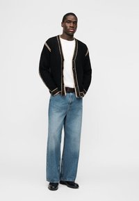 Άνδρας στέκεται με τα χέρια στις τσέπες, φορώντας μαύρη ζακέτα με μπεζ περίγραμμα, λευκό πουκάμισο, μπλε τζιν με φαρδιά μπατζάκια και μαύρα παπούτσια.