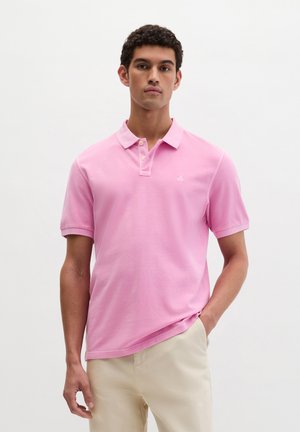 Mężczyzna w różowej koszulce polo z krótkim rękawem i beżowych spodniach stoi na tle jednolitego białego tła.