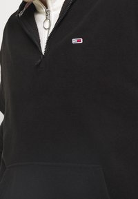 Svart fleece halvdragkedja med en broderad logotyp på vänster bröst, utrustad med en känguruficka och silverfärgad dragkedja.