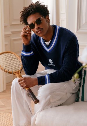 Un homme portant des lunettes de soleil et un pull bleu marine Wimbledon tient une raquette de tennis en bois vintage tout en étant assis à l'intérieur sur un canapé blanc.