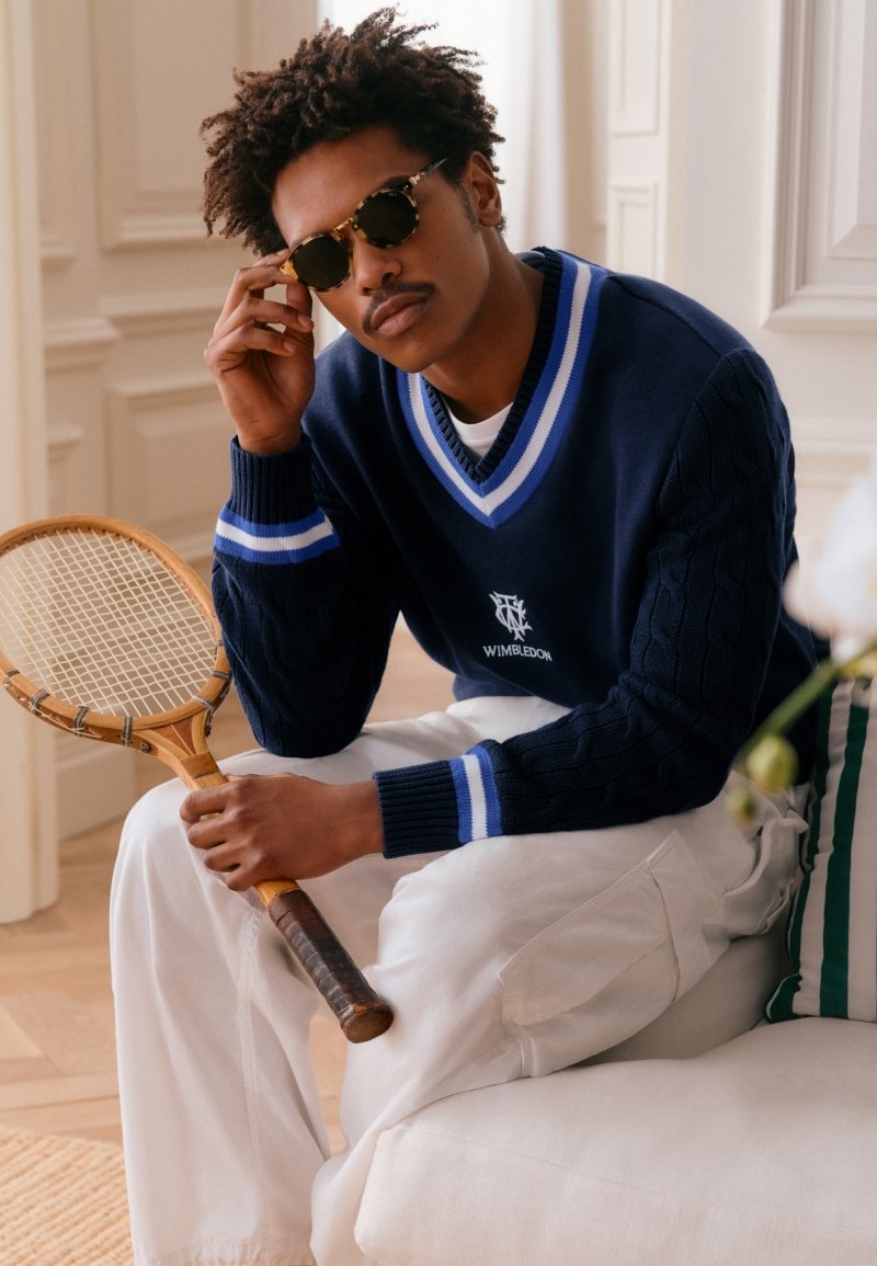 Man wearing sunglasses and a navy blue Wimbledon sweater holds a vintage wooden tennis racket while seated indoors on a white couch.