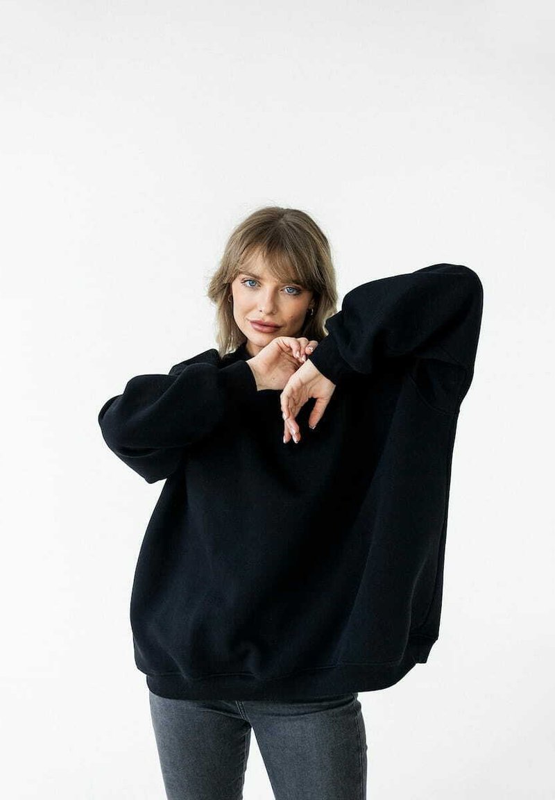Czarny, oversize sweter z długimi rękawami, rypsowymi mankietami i okrągłym dekoltem. Noszony z dopasowanymi, szarymi dżinsami. Gładka tekstura, minimalistyczny design.