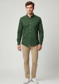 Camisa de botón verde oscuro de manga larga, combinada con chinos beige y zapatillas blancas. La camisa tiene un cuello y un pequeño detalle de logo.