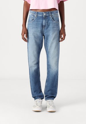 Person iført lyseblå slidte jeans, hvide sneakers og en cropped lyserød t-shirt, stående mod en ensartet hvid baggrund.