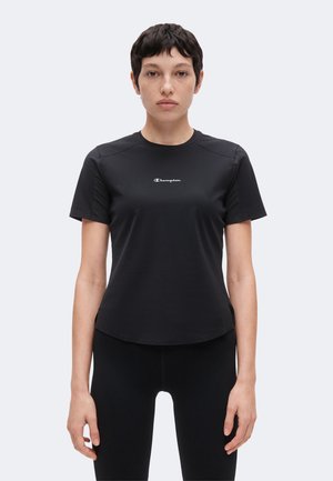 Person med kort mørkt hår iført en tætsiddende sort Champion t-shirt og sorte bukser, der står med fronten mod kameraet mod en ensfarvet baggrund.