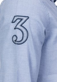 Vaaleansininen pitkähihainen paita, jossa vasemmassa hihassa tummansinisellä kirjailtu numero "3". Puuvillakangasta, sileä pinta.