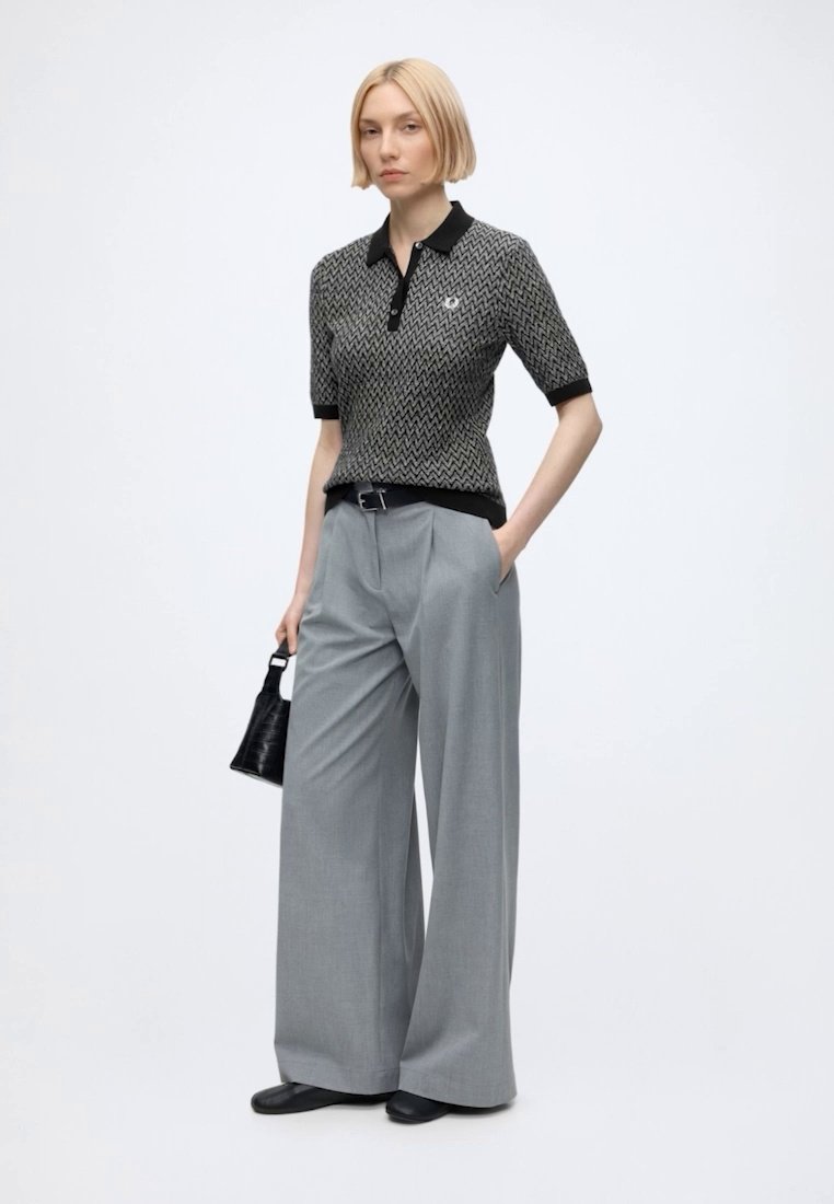 Femme blonde portant un polo noir et gris à motif zigzag, rentré dans un pantalon gris à jambes larges, tenant un petit sac à main noir, debout sur fond blanc.