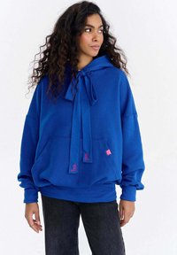 Hanorac albastru cu șnur la gât, buzunar frontal mare și manșete striate. Are accente roz cu logo și un croi lejer, supradimensionat.